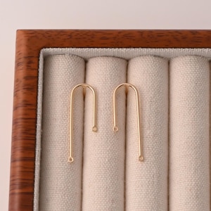 Op de afbeelding: Twee goudkleurige, U-vormige oorbelcomponenten worden gepresenteerd tegen een beige stoffen achtergrond in een houten sieradendoos. De oorbellen hebben een eenvoudig, minimalistisch ontwerp met kleine lussen aan de uiteinden.