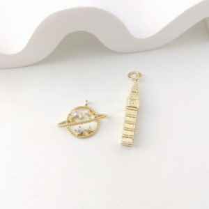 May include: Two gold-toned charms, one shaped like a planet with stars and a ring, and the other shaped like the Elizabeth Tower, also known as Big Ben, in London.