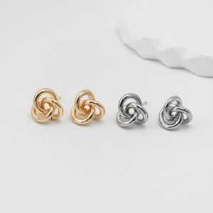 Puede incluir: Cuatro pares de pendientes de botón en tonos dorados y plateados. Cada pendiente presenta un diseño de nudo de tres bucles. Los pendientes se exhiben sobre una superficie blanca, mostrando su estética minimalista y moderna.