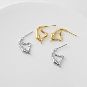 Pode incluir: São apresentados quatro brincos de formato abstrato, dois dourados e dois prateados. Cada brinco tem um pino para perfuração. Os brincos são apresentados sobre uma superfície branca. O design é moderno e minimalista.