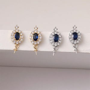 May include: Four oval-shaped pendant settings with a dark blue center stone, surrounded by small clear stones. Two are gold-toned, and two are silver-toned, each with a small loop and pin at the bottom for attaching a pendant.