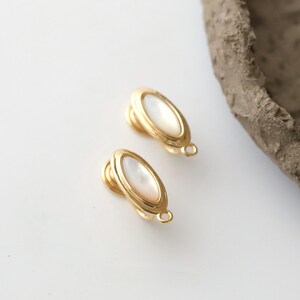 Pu&ograve; includere: Due orecchini a clip color oro con un centro ovale in madreperla. Gli orecchini hanno un design semplice ed elegante con un piccolo anello nella parte inferiore. Gli orecchini sono su una superficie bianca.