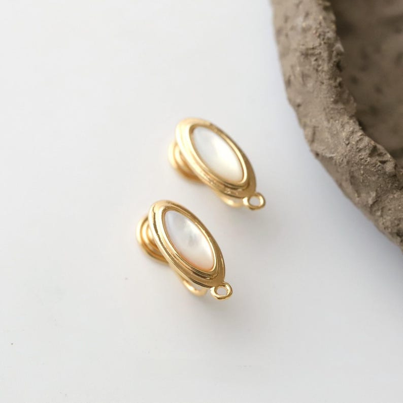 Pu&ograve; includere: Due orecchini a clip color oro con un centro ovale in madreperla. Gli orecchini hanno un design semplice ed elegante con un piccolo anello nella parte inferiore. Gli orecchini sono su una superficie bianca.