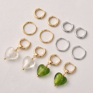 Op de afbeelding: Een set gouden en zilveren creolen met een paar gouden creolen met hartvormige bedels. De bedels zijn gemaakt van helder glas met zilveren vlokken en groen glas met gouden vlokken.