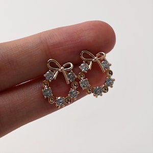 May include: A pair of gold-toned stud earrings. Each earring features a bow at the top and a circular design with clear, round gemstones. The earrings are set against a plain white background.