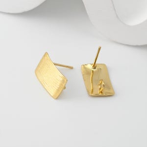 May include: Two gold-coloured stud earrings. Each earring has a rectangular back and a textured, angled front. The earrings are made of a shiny, metallic material. The posts are visible, and the earrings are set against a white background.