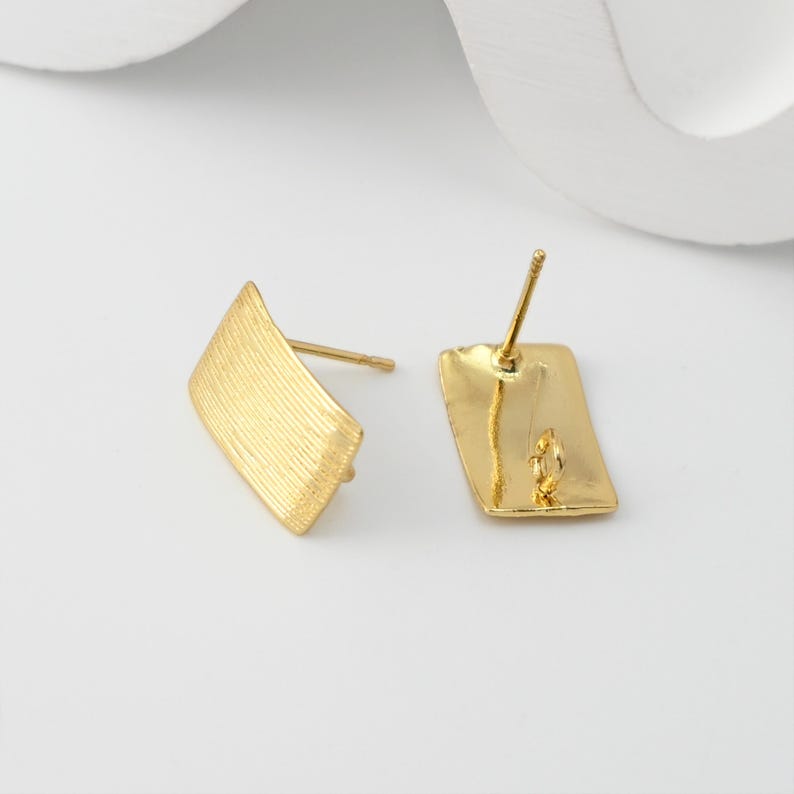 May include: Two gold-coloured stud earrings. Each earring has a rectangular back and a textured, angled front. The earrings are made of a shiny, metallic material. The posts are visible, and the earrings are set against a white background.