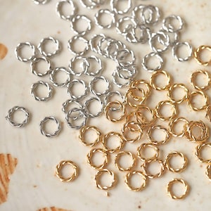 Puede incluir: Primer plano de una colección de pequeños anillos de salto de metal retorcido en tonos plateados y dorados. Estos componentes para la fabricación de joyas están dispersos sobre una superficie clara, mostrando su intrincado diseño.