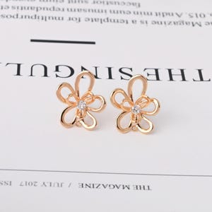 以下が含まれることがあります： ローズゴールド調の金属製スタッドピアスで、透かし彫りの花のような形をしています。それぞれのピアスの中央には、細い金属ワイヤーで形作られた花びらの中に、透明なラウンドカットの宝石がセットされています。