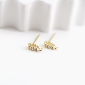 Könnte beinhalten: Zwei goldfarbene Ohrring-Verschlüsse mit jeweils einem kleinen, klaren Kristall-Akzent. Die Verschlüsse haben oben eine Öse und unten einen geraden Stift.