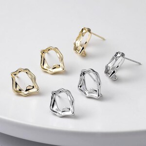 May include: A collection of gold and silver-toned stud earrings. The earrings have an irregular, organic shape with a hollow center. The earrings are displayed on a white surface, showcasing their reflective qualities and design.