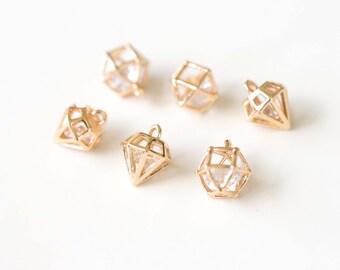 May include: Six gold-colored diamond-shaped charms with clear, faceted centers. The charms have a geometric cage design and small loops for attaching to jewelry. The charms are scattered on a white surface.