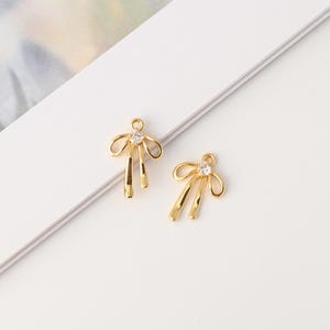 May include: Two gold-toned metal charms shaped like bows, each featuring a small, clear, round faceted stone set at the center. The charms have a smooth, polished finish and include a small circular loop at the top for attachment purposes.