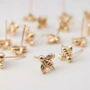 May include: A close-up of a group of gold bee stud earrings. The earrings are scattered on a white surface.