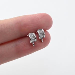 Puede incluir: Un par de pendientes de botón plateados. Cada pendiente presenta un diseño rectangular con múltiples piedras rectangulares pequeñas y transparentes. Los pendientes tienen un poste para la inserción y un pequeño lazo en la parte inferior.