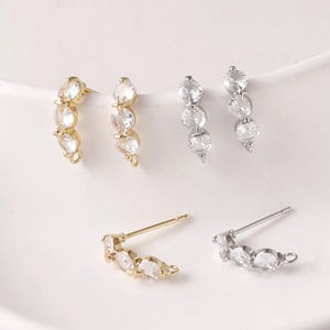 Könnte beinhalten: Ohrstecker in Gold und Silber, jeweils mit einer gebogenen Reihe klarer, rund geschliffener Zirkonia-Steine. Die Ohrringe haben ein minimalistisches Design. Die Ohrringe werden auf einer weißen Oberfläche präsentiert.