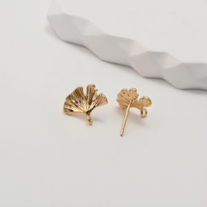 Puede incluir: Dos pendientes dorados con forma de hojas de ginkgo. Están sobre una superficie blanca con una forma geométrica blanca en el fondo. Los pendientes tienen un diseño detallado y texturizado.