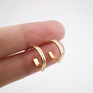 Può includere: Un paio di orecchini a cerchio in oro con un design delicato leggermente curvo. Gli anelli hanno una finitura liscia e lucida e presentano un piccolo ciondolo pendente nella parte inferiore.