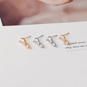 Pode incluir: Quatro brincos de pino dispostos em fila, com duas armações de metal em tom dourado e duas em tom prateado. Cada brinco possui um cristal transparente em forma de pera e um cristal redondo menor, com um pequeno elo metálico na base.