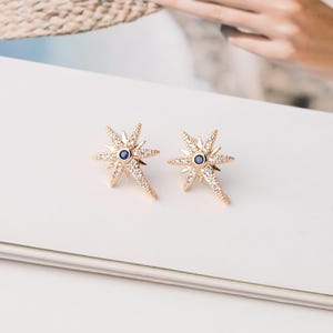 Pode incluir: Um par de brincos dourados em forma de estrela com uma pedra azul redonda central rodeada por pequenos cristais transparentes cravados em pavé. Os brincos têm um design de múltiplas pontas com um raio alongado estendendo-se para baixo.