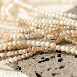 Peut inclure: Un brin de perles de coquillage irisées blanches et beiges. Les perles sont rondes et ont une finition lisse et polie. Les perles sont disposées en un tas lâche sur une surface texturée.