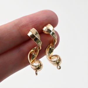 以下が含まれることがあります： リボンのようなデザインのゴールドカラーのイヤリングのペア。各イヤリングは、取り付け用の小さなループが付いた、ねじれた渦巻きの形をしています。イヤリングは光沢のある反射面で、ニュートラルな背景に設定されています。