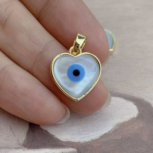 Könnte beinhalten: Goldener herzförmiger Anhänger mit einem blauen bösen Auge-Anhänger im Inneren. Der Anhänger ist aus Perlmutt.
