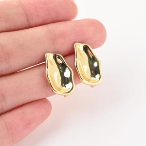Pode incluir: Um par de brincos de tarracha em tom dourado com um formato irregular, orgânico e semelhante a metal fundido, com uma superfície lisa e reflexiva. Os brincos têm um acabamento polido que capta a luz, criando brilhos intensos nos seus contornos curvos e irregulares.