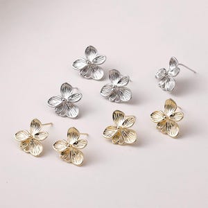 Pode incluir: Uma coleção de brincos de pressão em forma de flor em tons de prata e ouro. Os brincos apresentam um design delicado e vazado com cinco pétalas. Os brincos são exibidos em uma superfície branca, mostrando seus detalhes intrincados e brilho metálico.