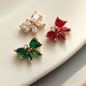 Peut inclure: Trois breloques en métal doré, chacune avec un motif floral. Une breloque présente des cristaux clairs en forme de cœur et ovales. Une autre a des cristaux rouges en forme de cœur, et la troisième a des cristaux verts en forme de cœur et ovales. Les breloques sont sur une surface blanche.