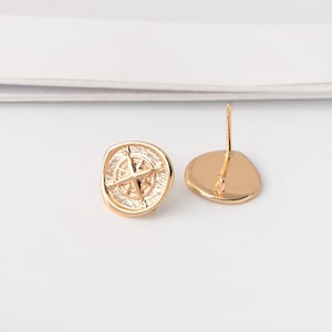 Peut inclure: Une paire de boucles d'oreilles dorées. Chaque boucle d'oreille présente un motif de rose des vents dans un cadre circulaire. Les boucles d'oreilles ont une tige et un fermoir pour un port sûr. Le design est détaillé, sur fond blanc.