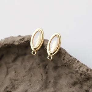 Pu&ograve; includere: Due orecchini a lobo ovali color oro con un centro perlaceo bianco sporco. Gli orecchini hanno un design semplice ed elegante, con un piccolo anello nella parte inferiore per il fissaggio. Gli orecchini sono esposti su una superficie grigia strutturata.