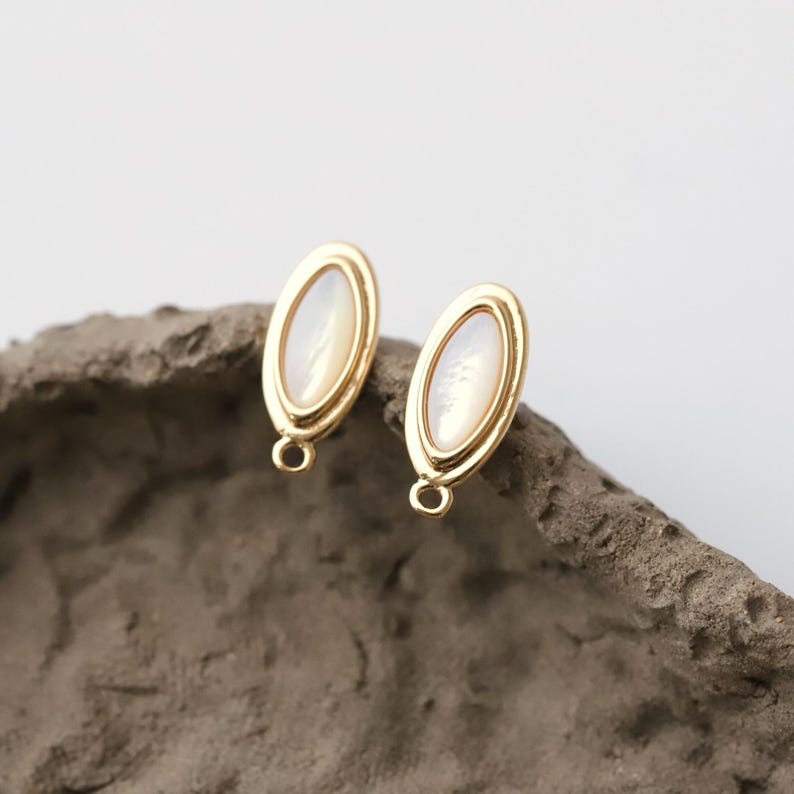 Pu&ograve; includere: Due orecchini a lobo ovali color oro con un centro perlaceo bianco sporco. Gli orecchini hanno un design semplice ed elegante, con un piccolo anello nella parte inferiore per il fissaggio. Gli orecchini sono esposti su una superficie grigia strutturata.