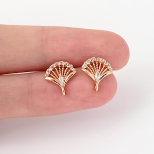 May include: A pair of gold-coloured fan-shaped stud earrings. Each earring has a row of small, clear crystals along the top edge and a larger, teardrop-shaped crystal in the centre. The earrings are held in a person's hand.