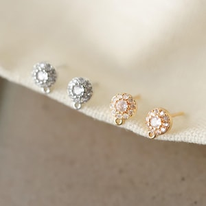 Puede incluir: Primer plano de cuatro pendientes de botón. Dos son plateados con piedras transparentes y dos son dorados con piedras rosas. Cada pendiente tiene un pequeño lazo en la parte inferior. Los pendientes se muestran sobre una tela blanca.