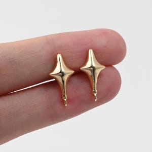 Pode incluir: Dois brincos dourados em forma de estrela. Cada brinco apresenta um design de estrela de quatro pontas com uma base curva e alongada e um pequeno laço na parte inferior. Fundo neutro.