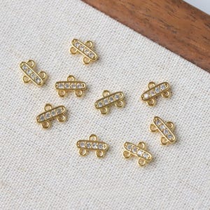 Puede incluir: Primer plano de varios conectores de joyería pequeños, de color dorado. Cada conector presenta una barra rectangular con una fila de piedras claras y brillantes. Los conectores tienen pequeños bucles en cada extremo para sujetar.