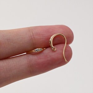 May include: Close-up of gold-colored earring components. One is a small, elongated piece with embedded clear stones. The other is a curved ear wire with a small loop at the end. The components are for jewelry making.