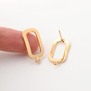 May include: Two gold-colored rectangular earring components. One is held by a finger, the other is a stud earring. Each has a small loop at the bottom and a post for attachment. The earrings are on a white background.