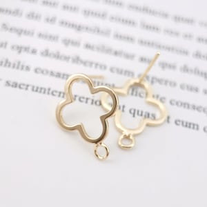 Op de afbeelding: Een paar goudkleurige klavervormige oorstekers. Elke oorbel heeft een klaverontwerp met vier bladeren, een pin voor piercing en een kleine lus aan de onderkant. De oorbellen staan tegen een vage achtergrond.