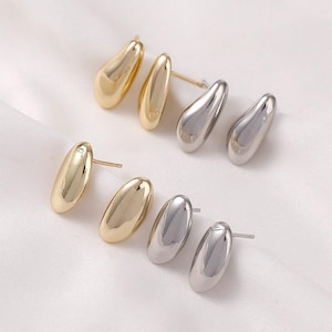 Puede incluir: Seis pares de pendientes de botón con forma de lágrima. Tres pares son dorados y tres pares plateados. Los pendientes son lisos y brillantes, con un diseño minimalista. Se muestran sobre una superficie blanca.