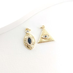Puede incluir: Dos colgantes de tono dorado con amuletos de ojo turco azul. Un colgante tiene forma triangular con un diseño de rayos de sol. El otro colgante tiene forma ovalada con un diseño detallado.