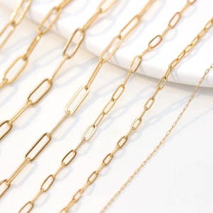 Può includere: Cinque catene d'oro di diverse lunghezze e spessori, tutte realizzate con un design semplice e a maglie. Le catene sono disposte su una superficie bianca.