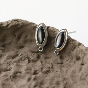 Pu&ograve; includere: Un paio di orecchini a lobo ovali con una pietra centrale scura e iridescente incastonata in una montatura in metallo color argento. Ogni orecchino ha un piccolo anello in basso e un perno.