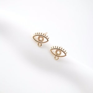 Könnte beinhalten: Ein Paar goldfarbene Ohrringe mit einem Augen-Anhänger und einer kleinen Schlaufe unten. Die Ohrringe haben die Form eines Auges mit Wimpern.