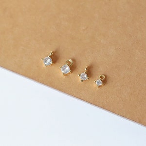 May include: Four small gold-toned jewellery charms, each featuring a single round clear stone set in a four-prong mount with a small circular loop at the top for attachment. The charms vary slightly in size, arranged in a row from largest to smallest.