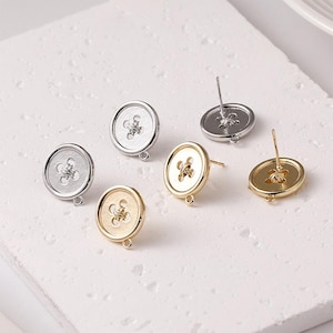 Puede incluir: Pendientes de botón en tonos plateados y dorados. Los pendientes presentan un diseño floral detallado en el centro y un pequeño lazo en la parte inferior. Los postes son visibles en la parte posterior. Medidas en centímetros.