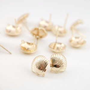 Könnte beinhalten: Ein Paar goldfarbene Ohrringe in Muschelform. Die Ohrringe liegen auf einer weißen Oberfläche.