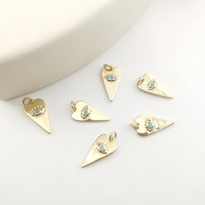 Puede incluir: Seis encantos de corazón de oro con un diseño de ojo malvado azul. Los encantos están sobre una superficie blanca.