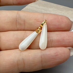 Puede incluir: Dos colgantes de color blanco en forma de lágrima con tapas doradas. Los colgantes están hechos de nácar y tienen un acabado liso y pulido.